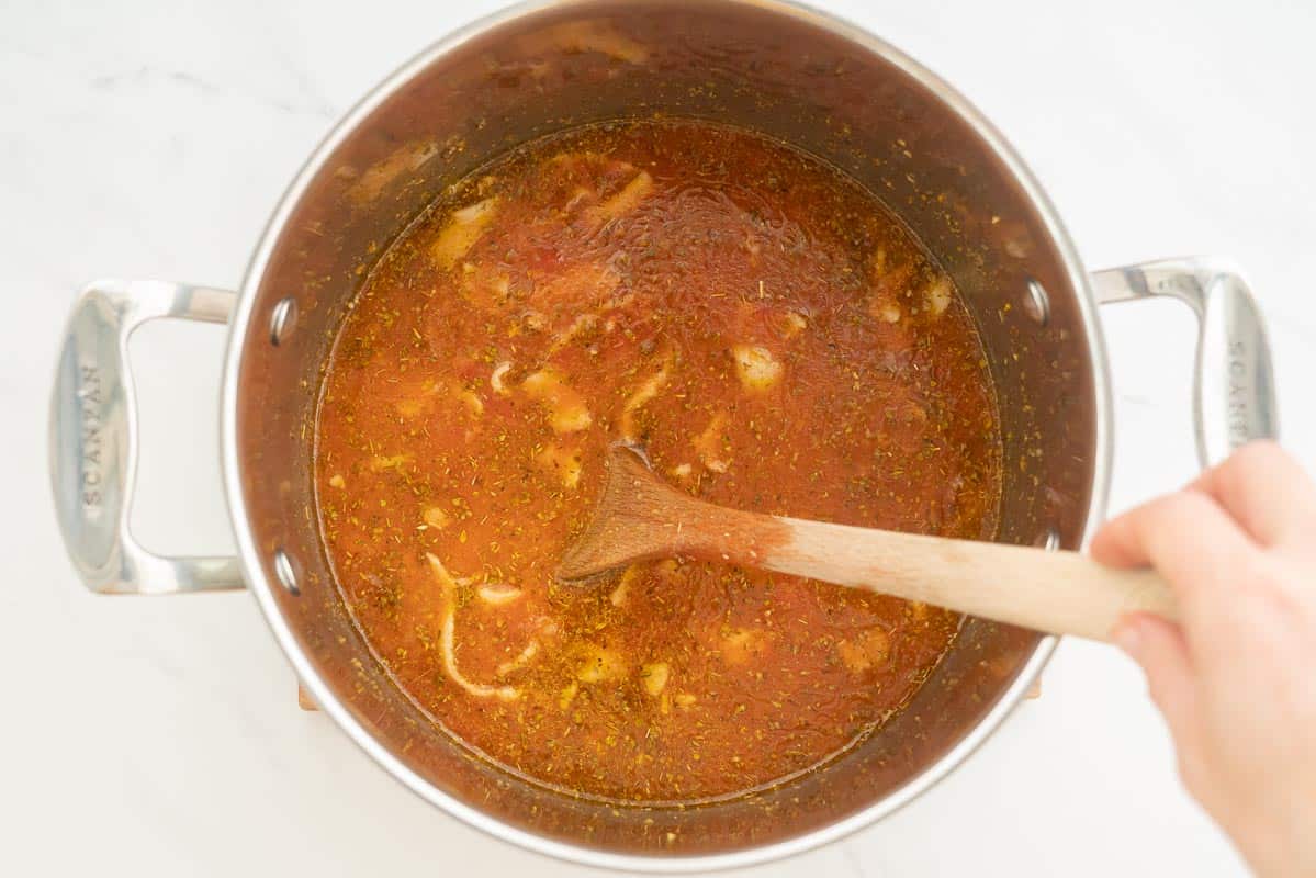 Tomato soup in a large saucepan being stirred with a wooden spoon.