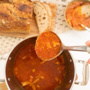 Tomato soup being ladled from a large saucepan into a bowl with crusty bread.