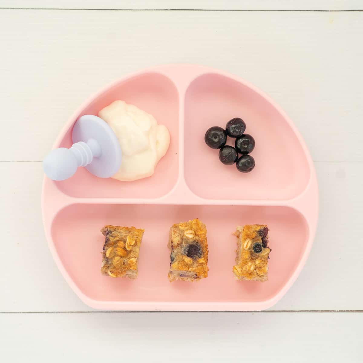 A pink divided baby plate with slices of baked oats, yoghurt and blueberries
