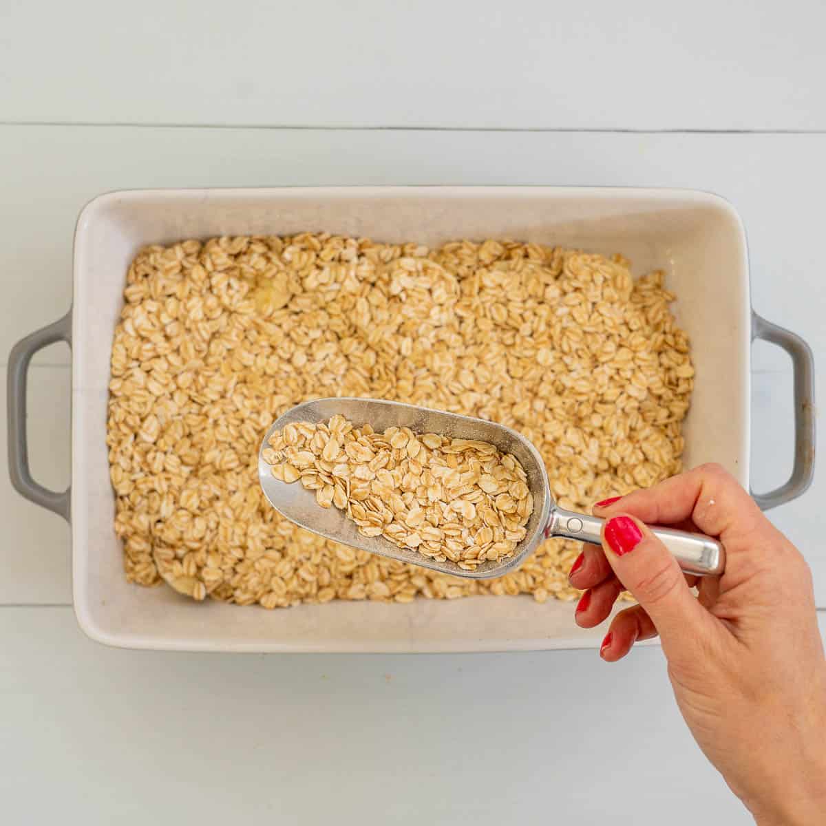 A woman's hand holding a scoop of rolled oats above a rectangular baking dish.