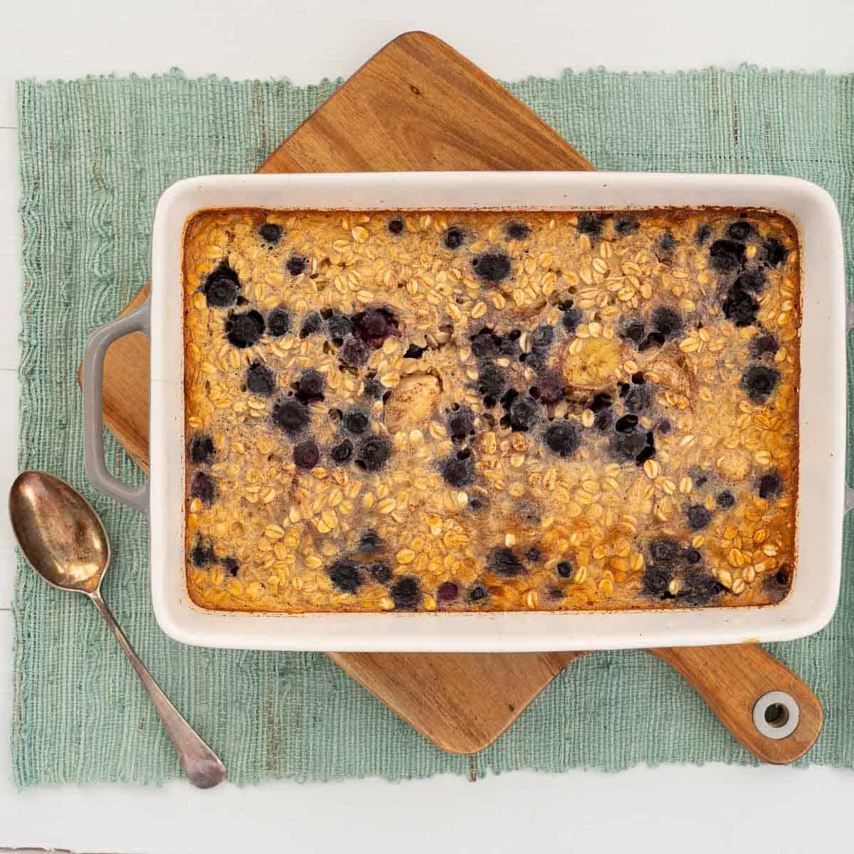A baking dish filled with blueberry baked oats sitting on a wooden board with a serving spoon.