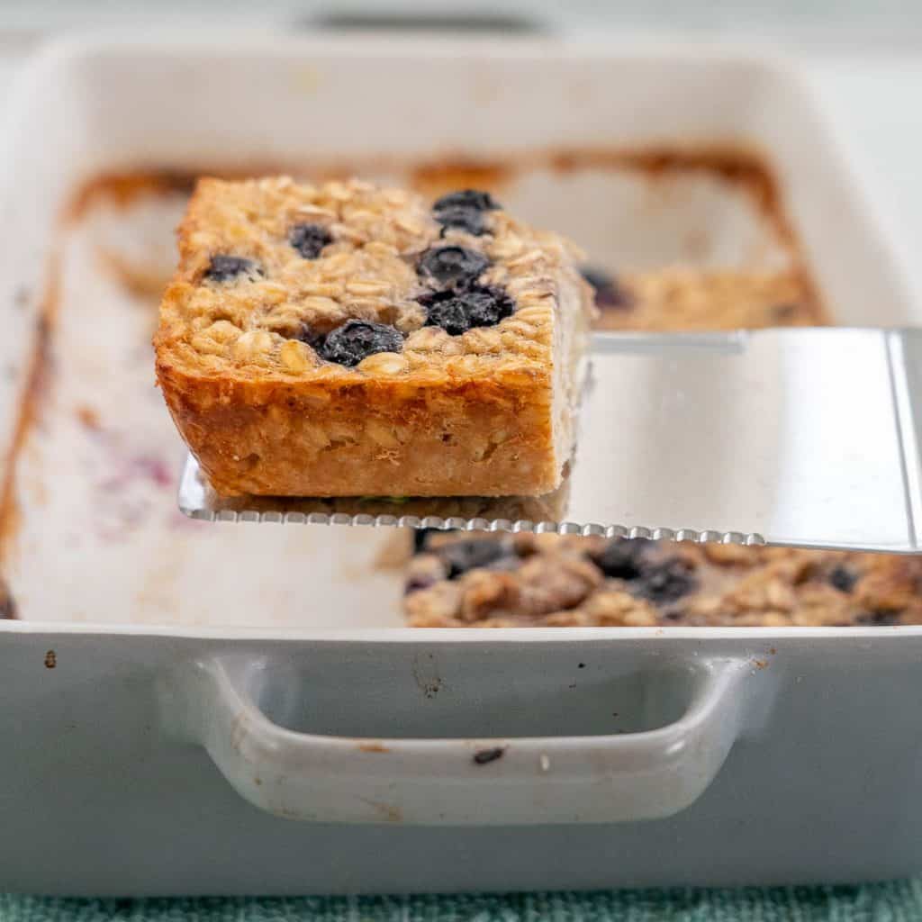 A square slice of baked oats topped with blueberries being lifted out of a baking dish.