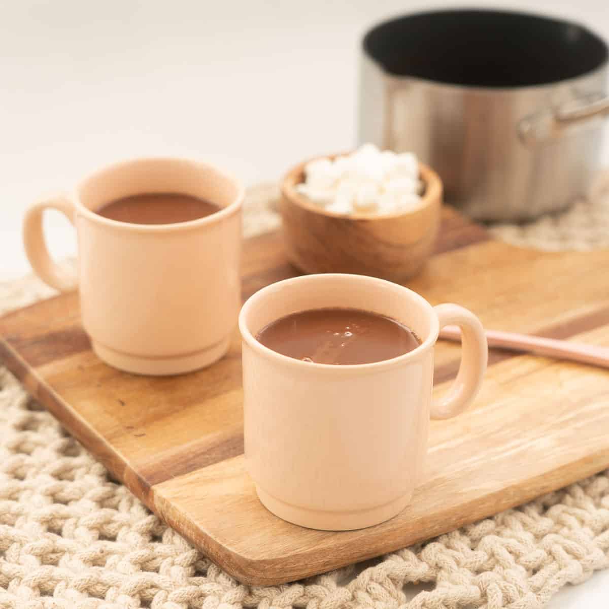 2 light pink mugs sitting on a wooden board filled with hot chocolate.