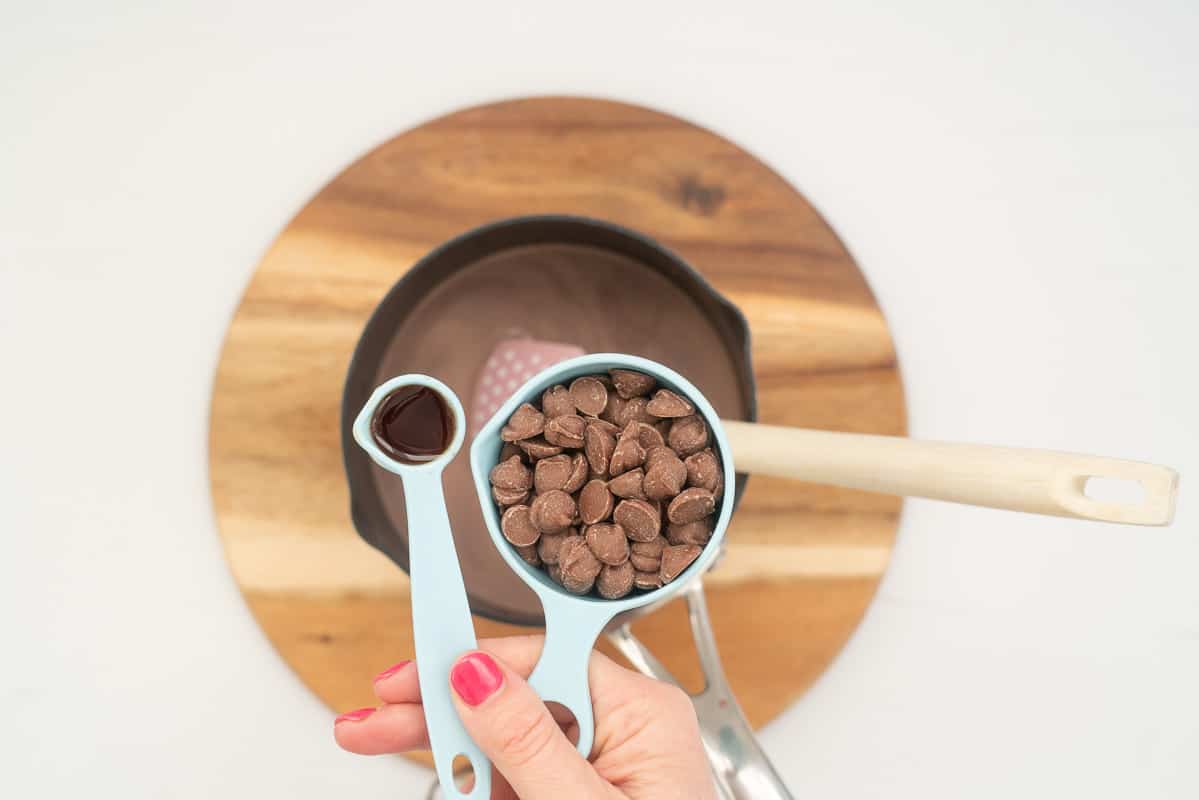 A woman's hand holding ½ a cup of chocolate drops and 1 teaspoon of vanilla essence above a saucepan of hot chocolate.