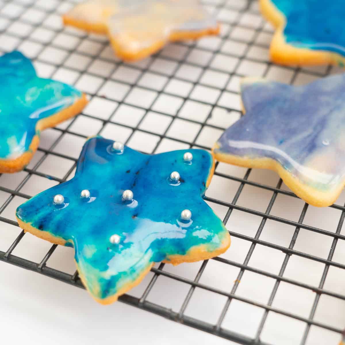 A blue galaxy iced cookie on a cooling rack decorated with edible silver balls.