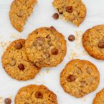 Chocolate chip oatmeal cookies lyung on white baking paper with oats and chocolate drops scattered around.