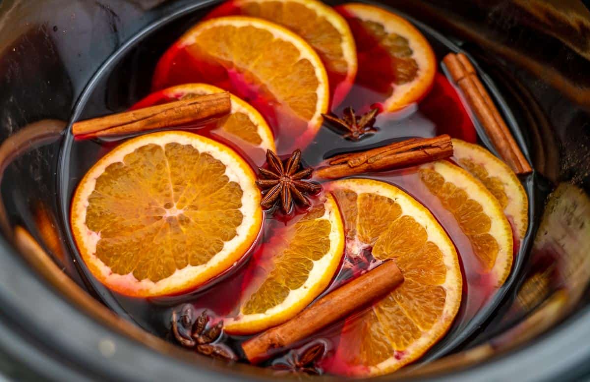 Dark grape juice in a slow cooker with cinnamon quills, star anise and orange slices floating on the surface.