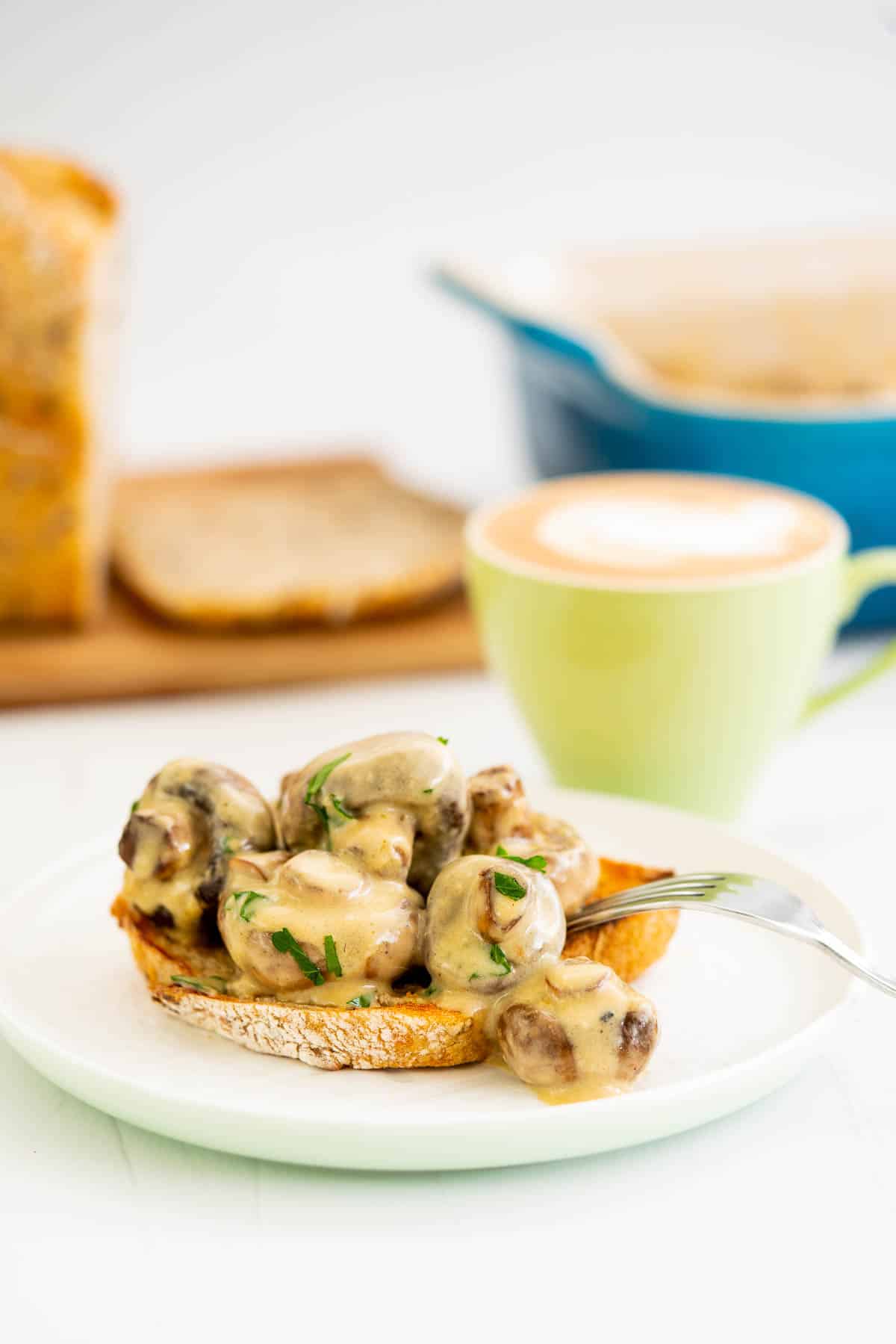Creamy roast mushrooms on grainy toast, a cup of coffee and loaf of bread in the background.