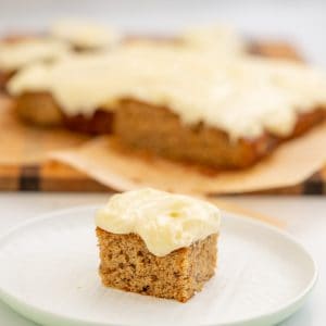 A square piece of banana cake frosted with cream cheese icing sitting on a white plate.
