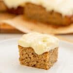A square piece of banana cake frosted with cream cheese icing sitting on a white plate.