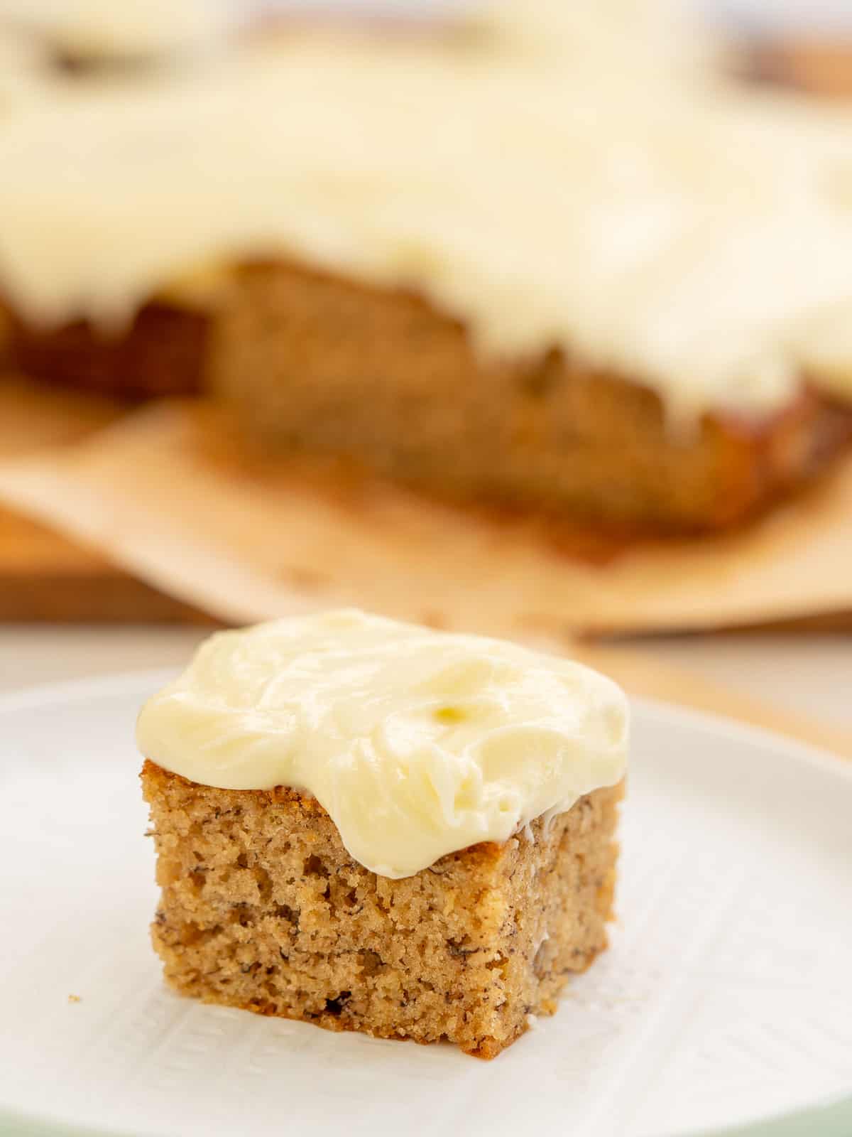 A square piece of banana cake frosted with cream cheese icing sitting on a white plate.
