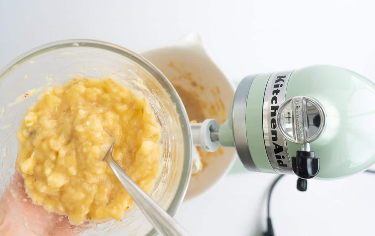 A glass bowl of mashed bananas being held above a stand mixer.