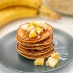 A stack of 5 pancakes on a blue plate with sliced banana and maple syrup.