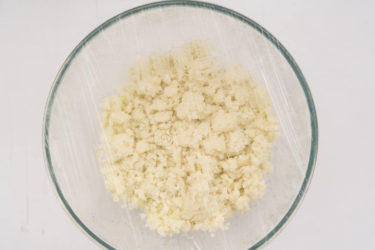 Riced cauliflower and onion steaming in a microwave proof bowl covered with cling film.