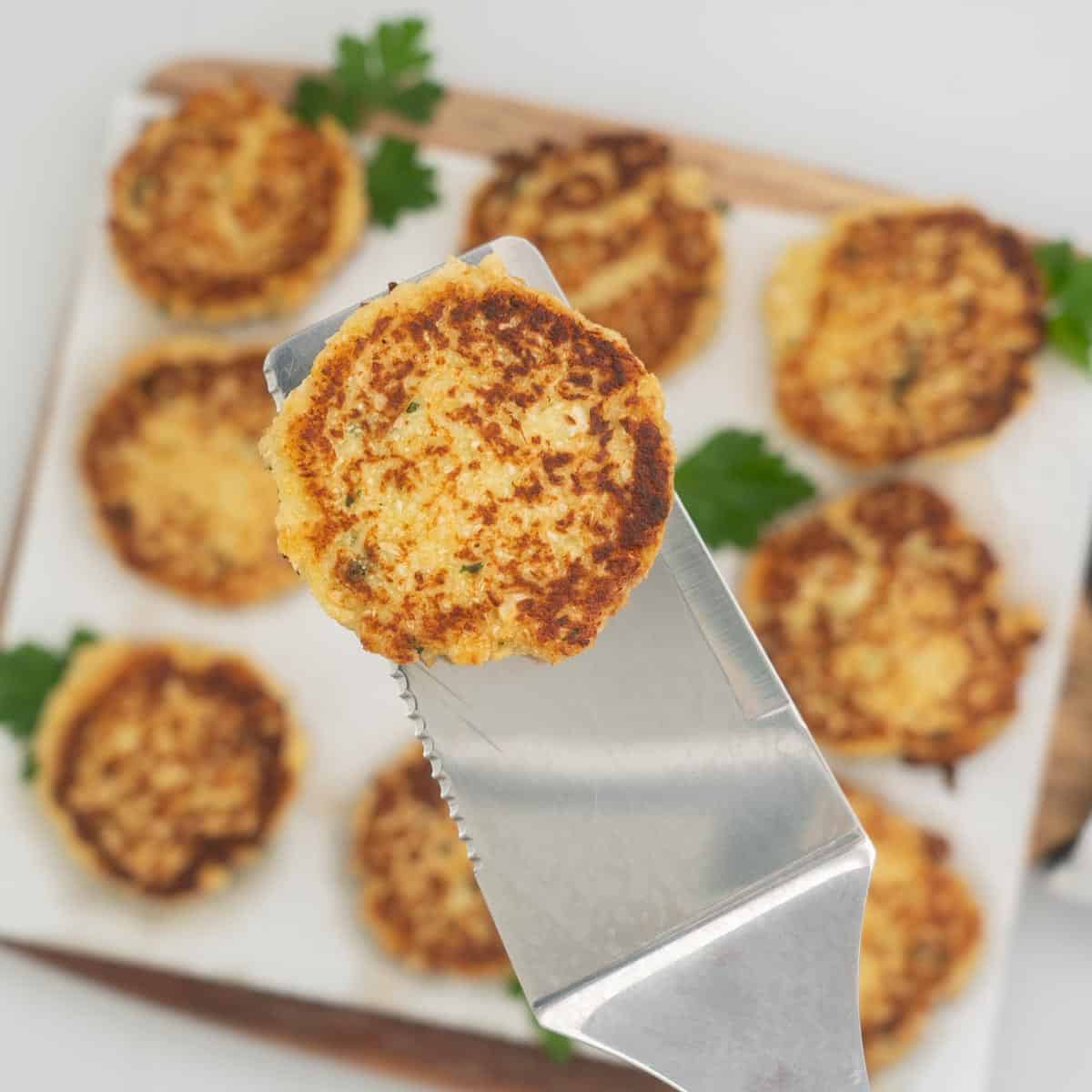 A cauliflower fritter on a stainless steel spatula being held above a platter of fritters.