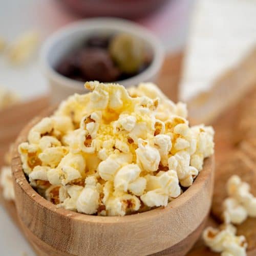 A small wooden bowl of cheese popcorn, antipasto platter ingredients in the background.
