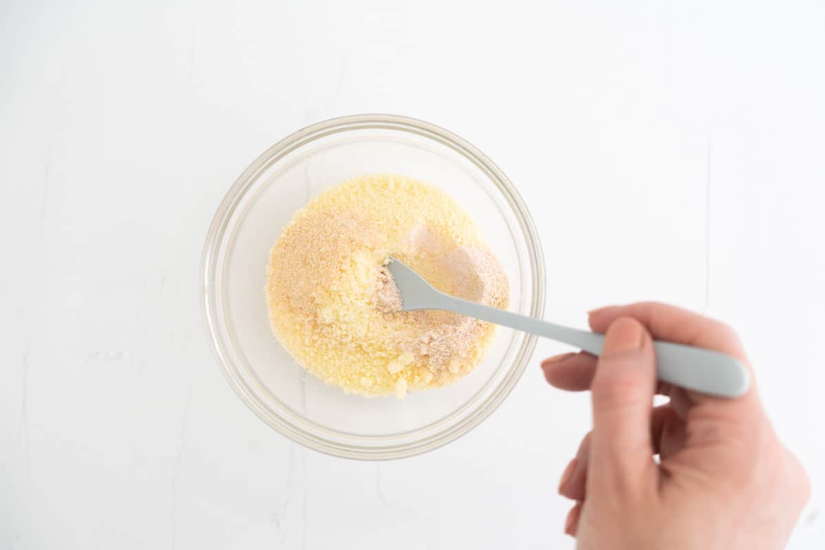 A woman's hand mixing powdered parmesan cheese and seasonings in a small glass bowl. 