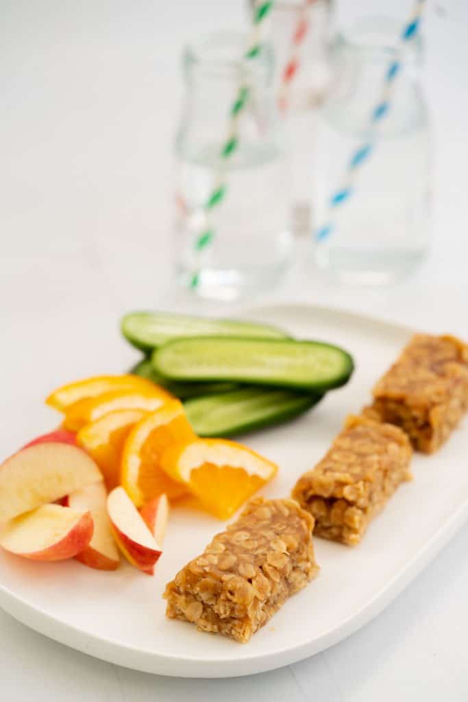 Three peanut butter bars on a white platter with cut up fruit and vegies.