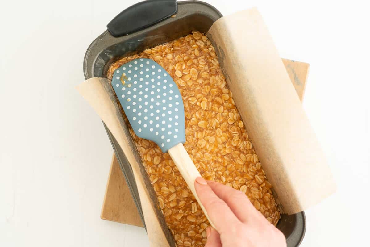 A blue spatula pressing oat bar mix into the base of a lined loaf tin.