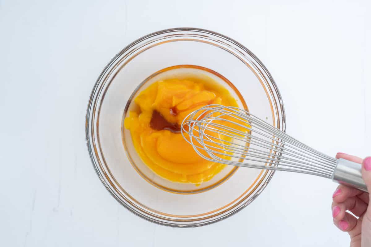 Pumpkin puree, oil, vanilla and maple syrup in a glass bowl, a whisk hovering above the bowl.