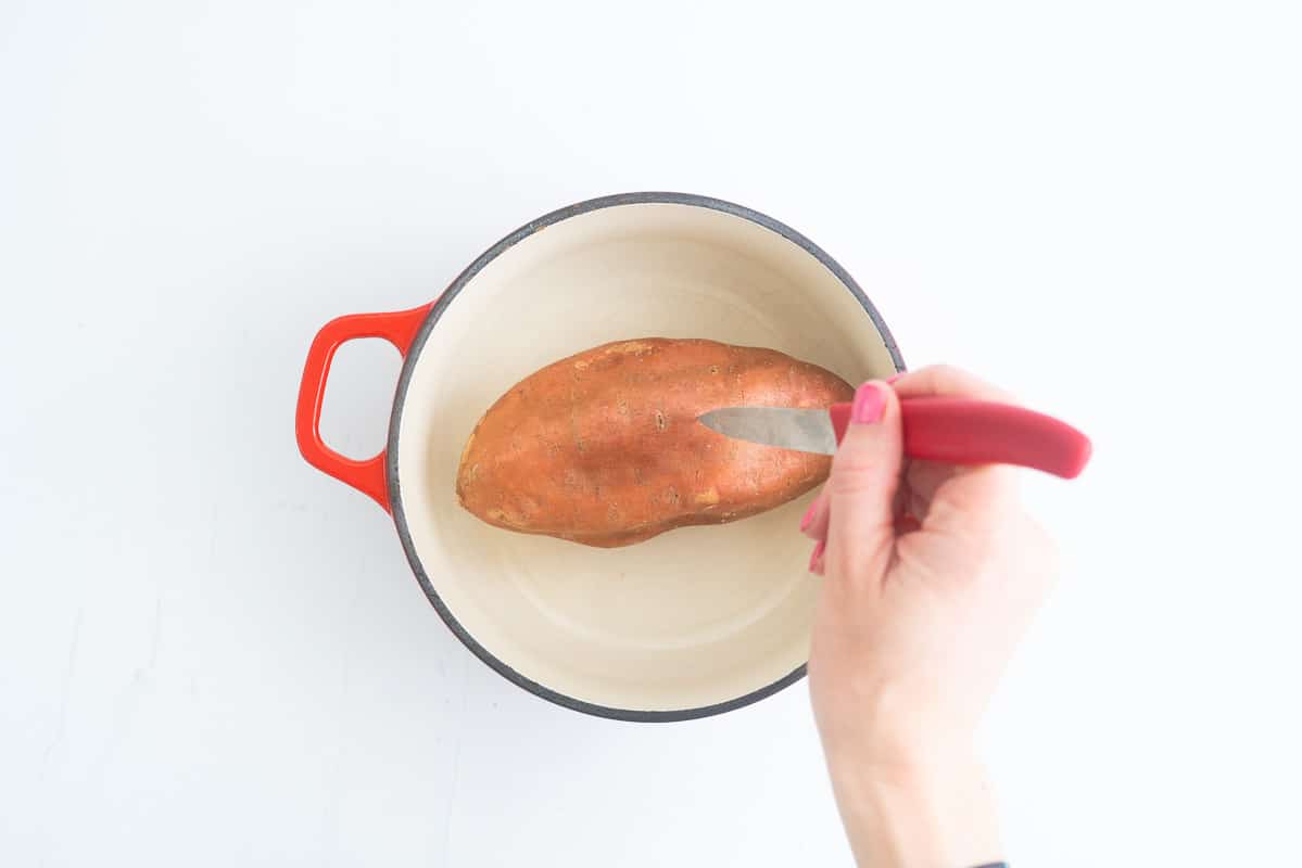 A sweet potato in an oven proof dish, a womans hand piercing the skin with a sharp knife.