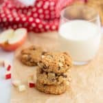 A stack of three oat meal cookies sitting on crinkled parchment paper with a glass of milk in the background.
