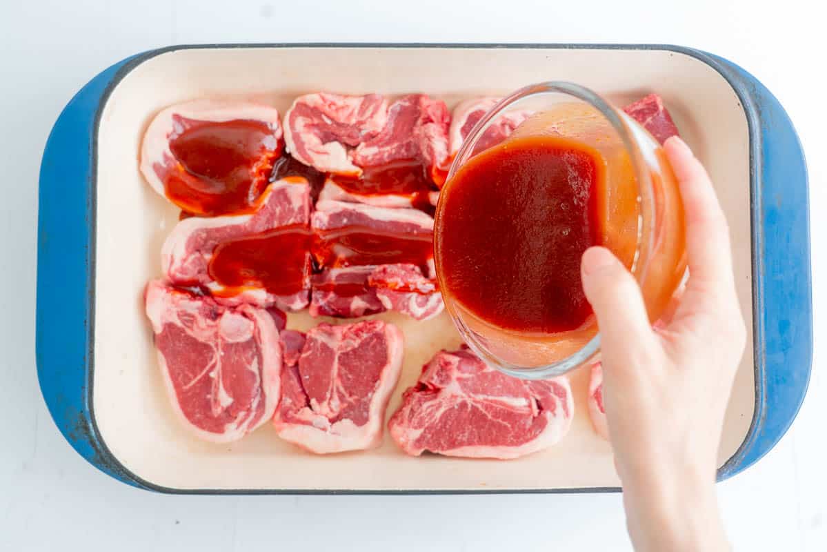 Red marinade being poured over lamb chops in a blue ceramic baking dish.