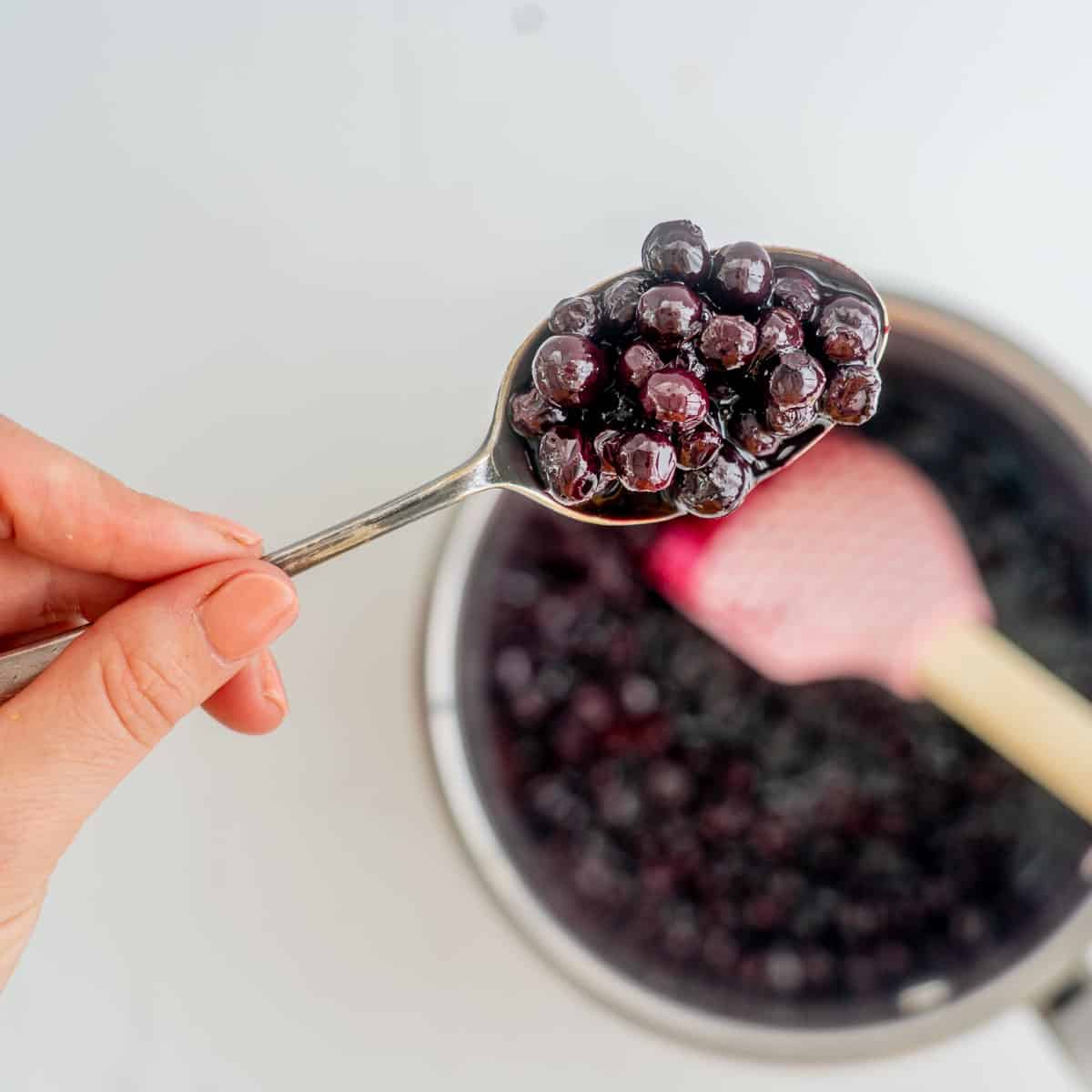 A spoonful of blueberry sauce held above a saucepan of blueberry compote.