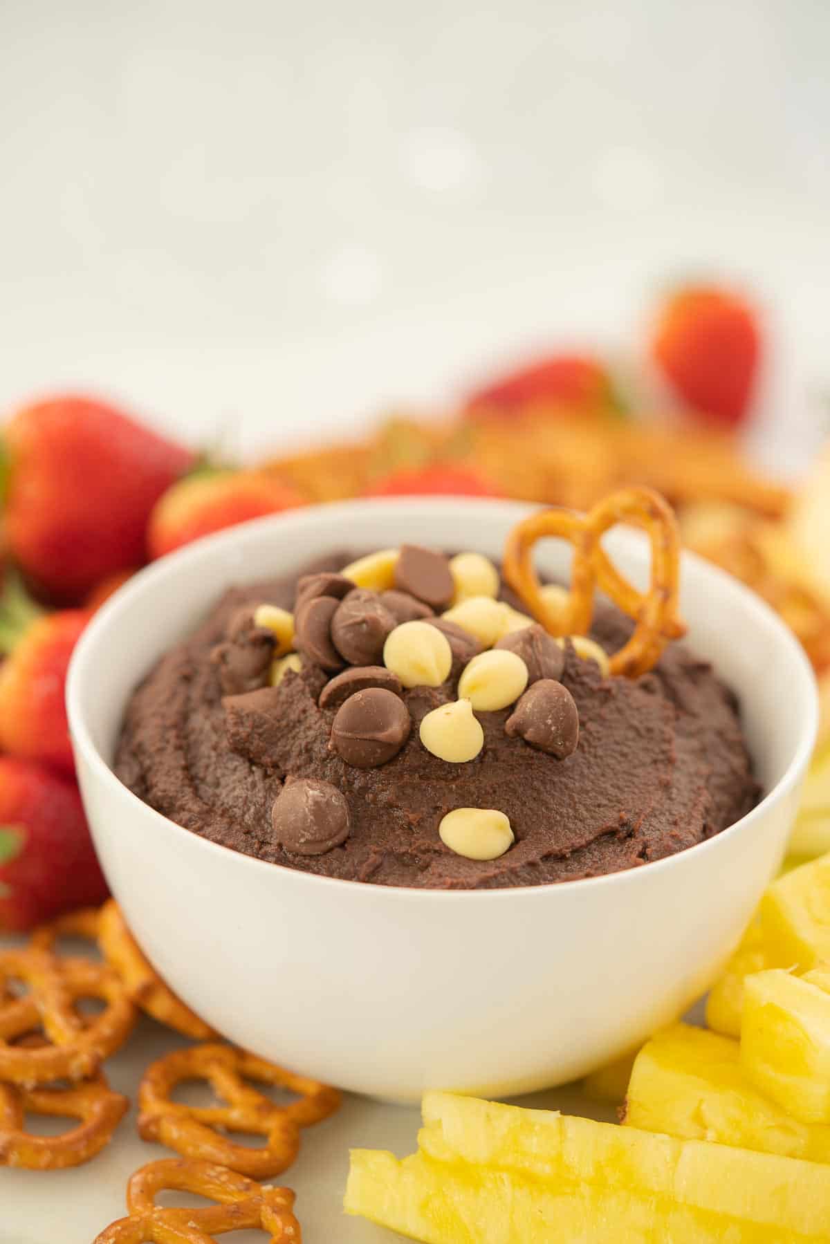 A bowl of chickpea brownie batter dip topped with chocolate drops and pretzels on a platter of fruit. 