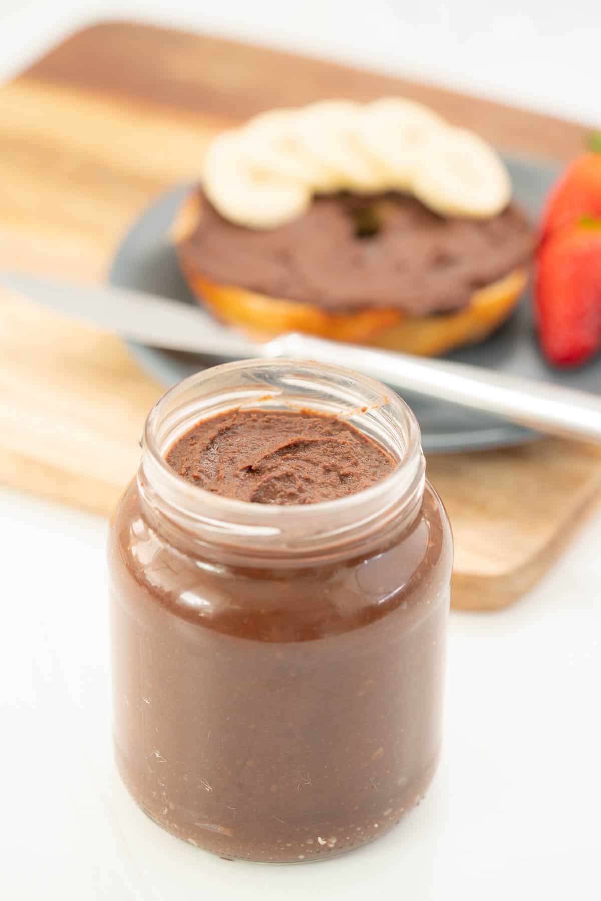 A jar of chocolate spread sitting in front of a bagel, plate and knife. 