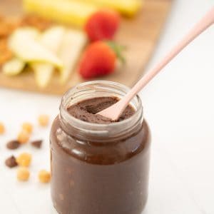 A jar of chocolate spread with a pink spoon, in front of a wooden board of fruit.
