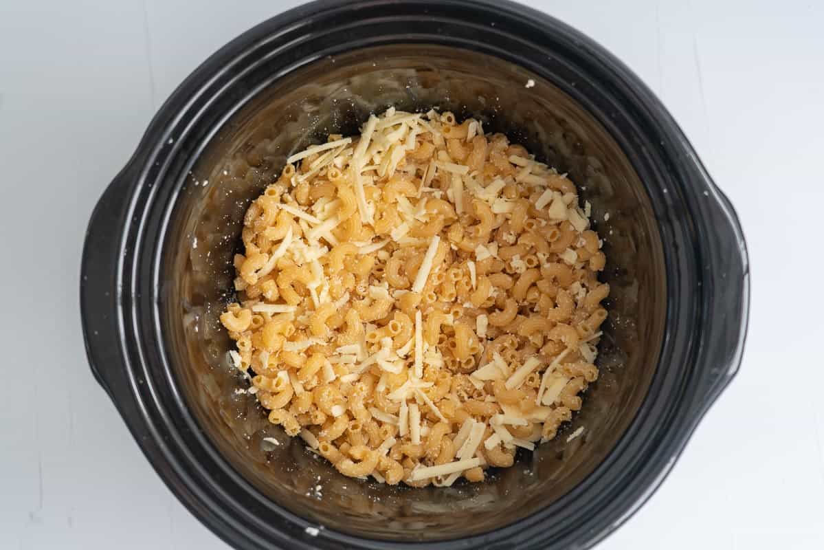 Dry macaroni elbow pasta and grated cheese in a black slow cooker dish.