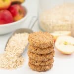 A stack of 5 oatmeal toddler cookies on a bench top with rolled oats and apples in the background.