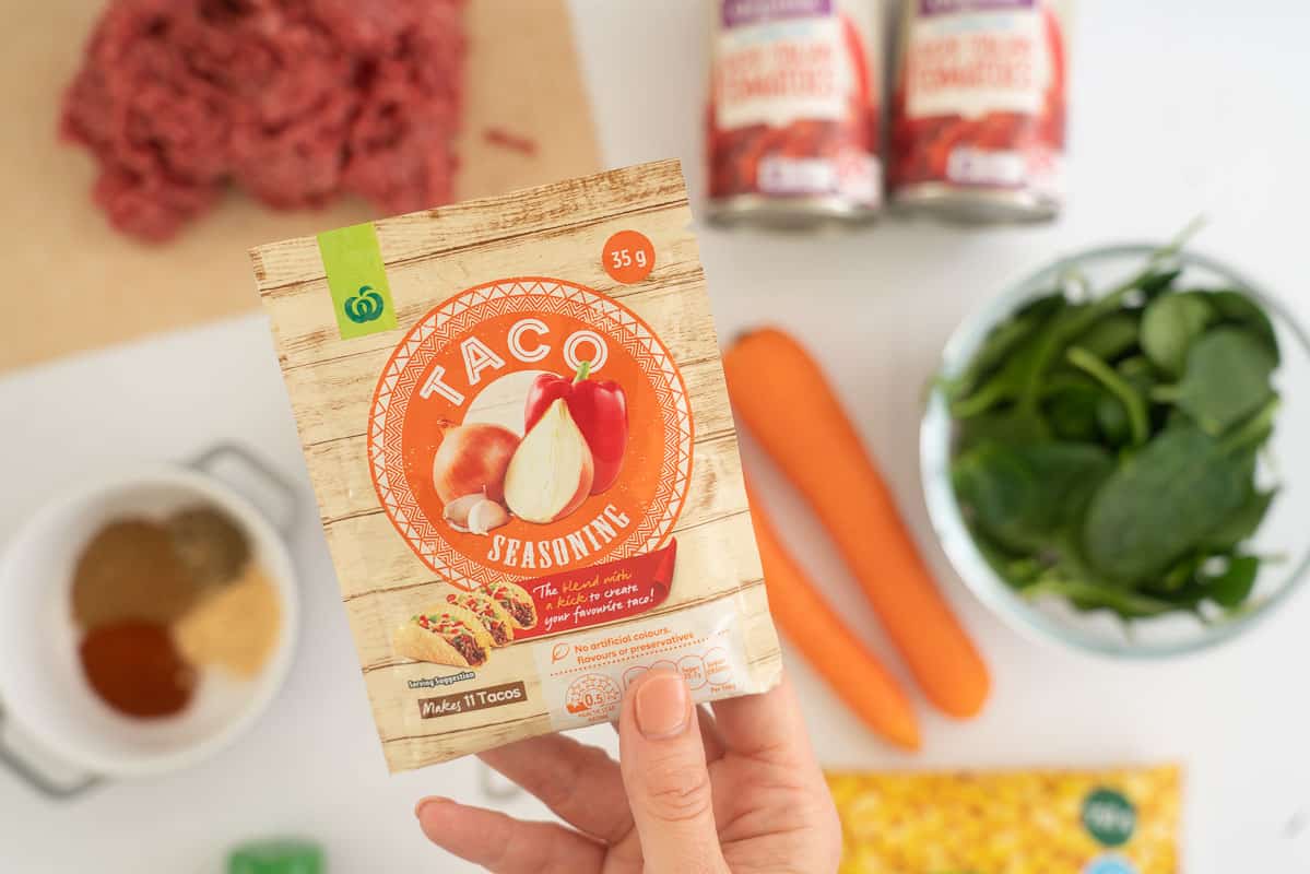 A hand holding a packet of taco seasoning above a bench of fresh produce.