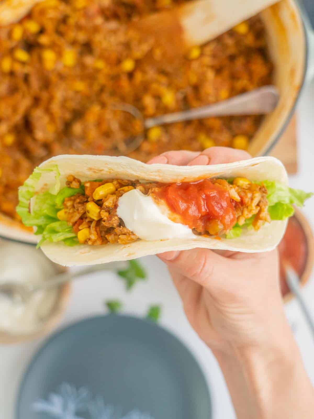 A hand holding a taco filled with mince, rice, corn, lettuce and salsa.