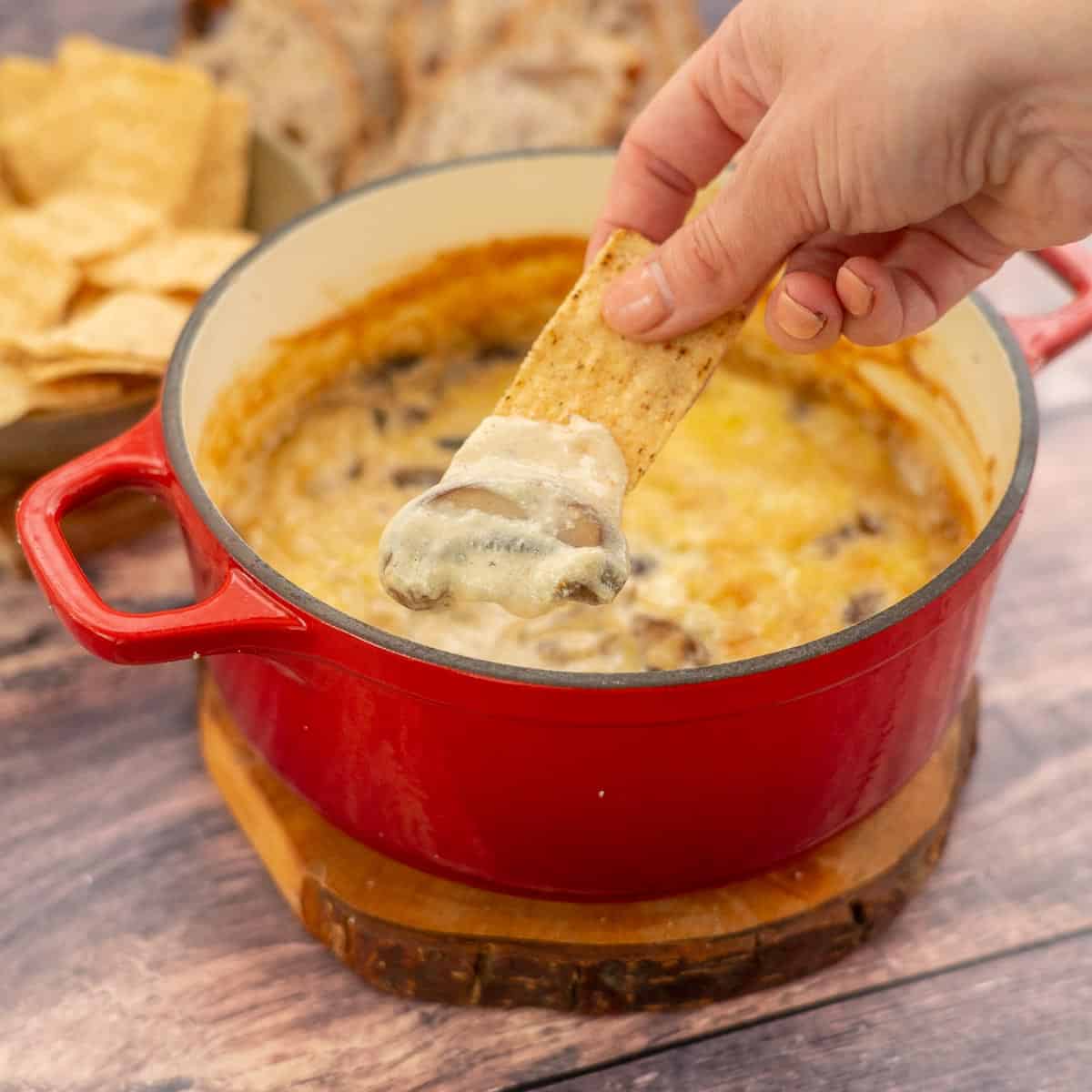 A hand scooping baked cheese dip fro a red dish with a corn tortilla chip.