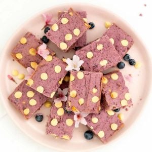 Purple cookie bars on a pink plate scattered with blueberries, pink flours and white chocolate chips.