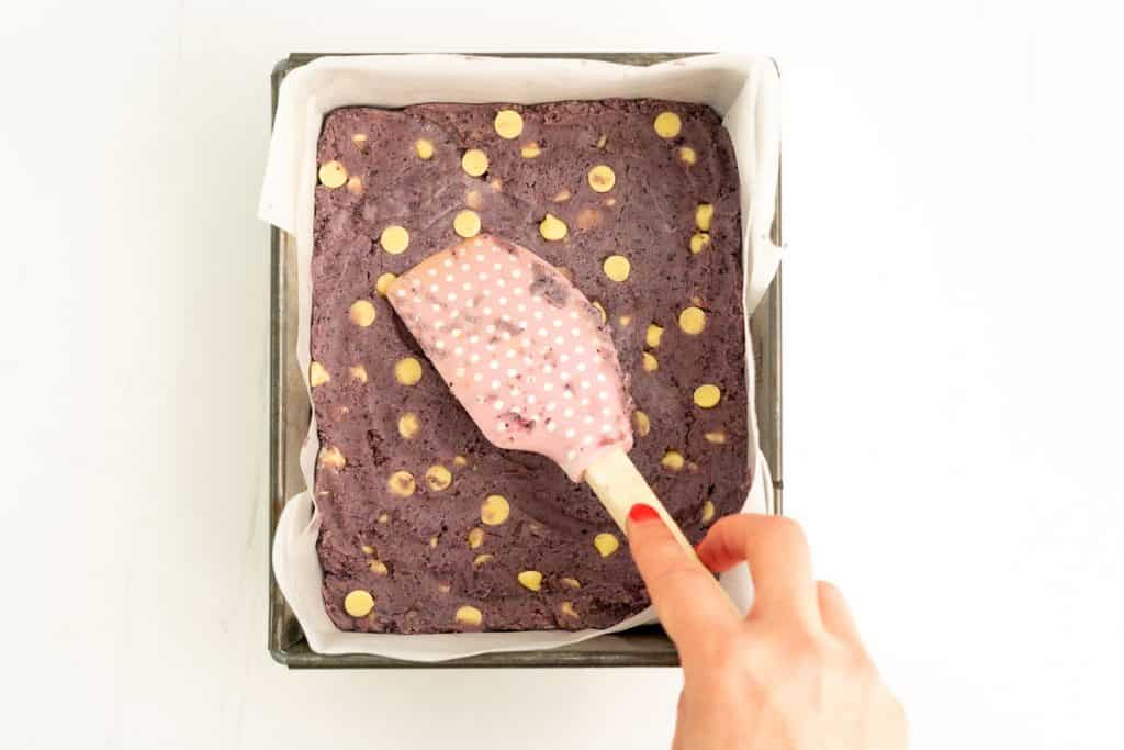 Purple cookie dough studded with white chocolate drops being pressed into the baae of a square cake tine with a pink polka dotted spatula.