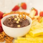 A bowl of chickpea brownie batter dip topped with chocolate drops and pretzels on a platter of fruit.