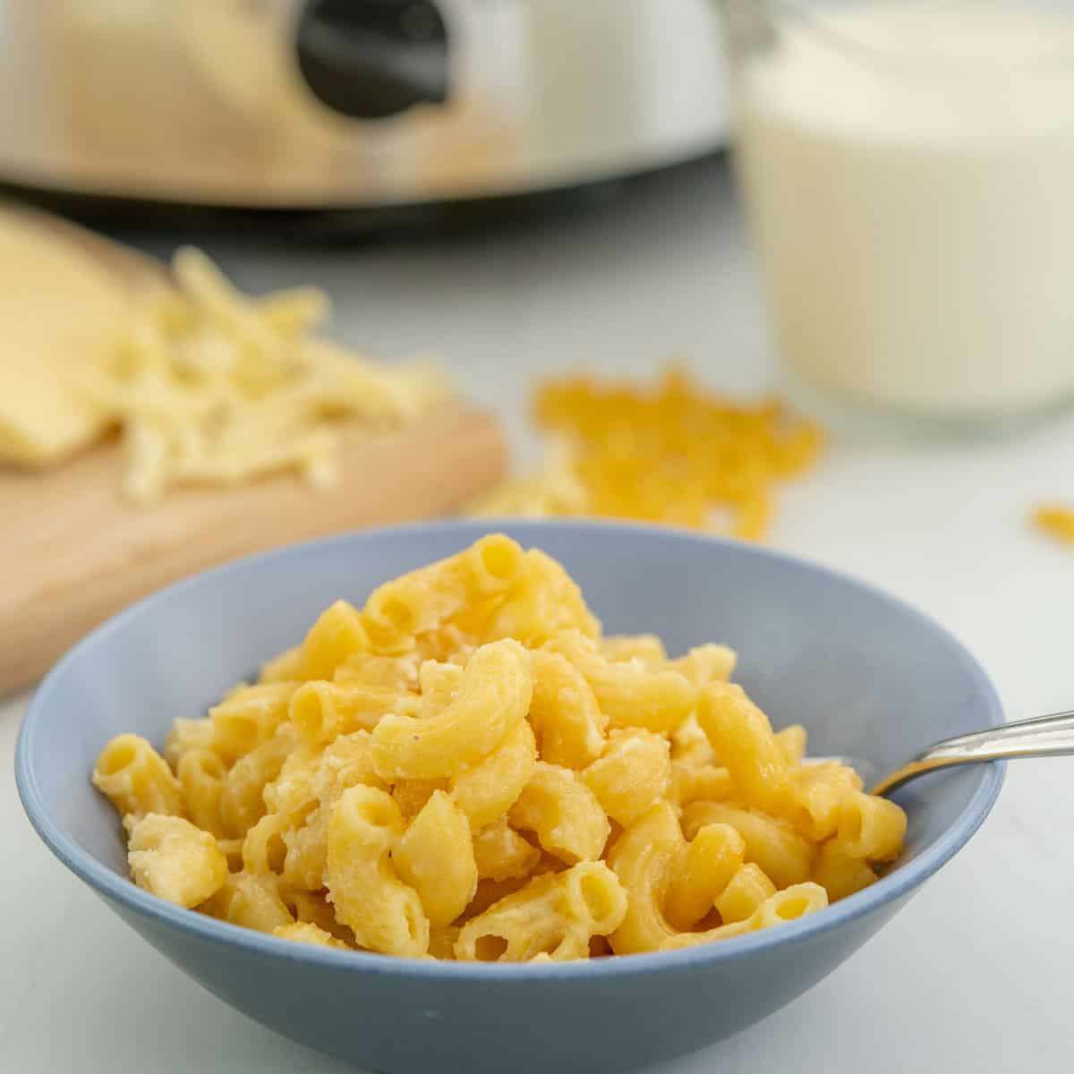 A two tone ceramic bowl of macaroni cheese sitting in front of ingredients and a slow cooker. 