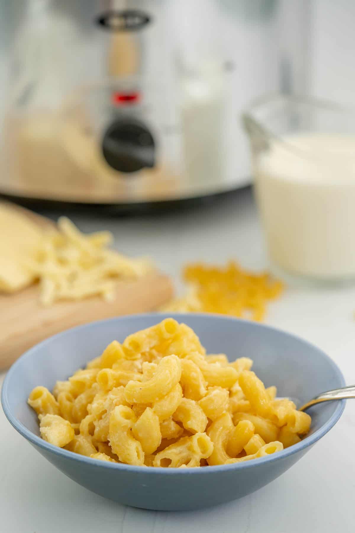 A two tone blue ceramic bowl of macaroni cheese in front of slow cooker, grated cheese and a glass jug of milk.