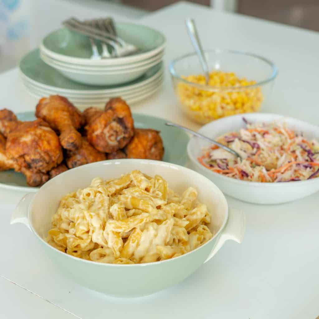 Cheese pasta, coleslaw, chicken drumsticks, and corn kernels on a table. 