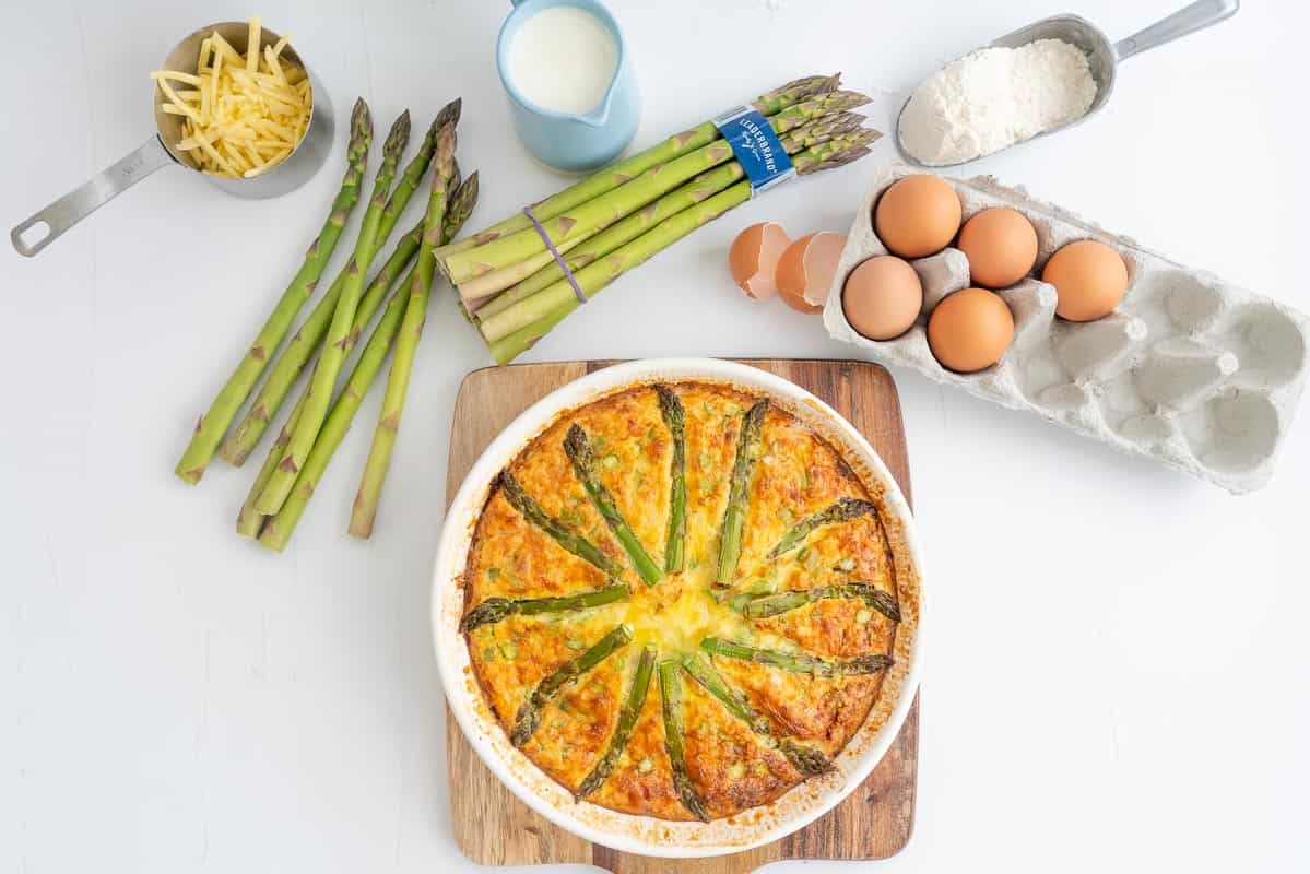 Golden brown cooked asparagus quiche on a wooden board surrounded by quiche ingredients.