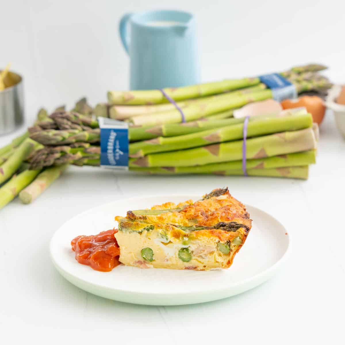 A slice of asparagus quiche on a white plate garnished with tomato relish, bunches of asparagus in the background.