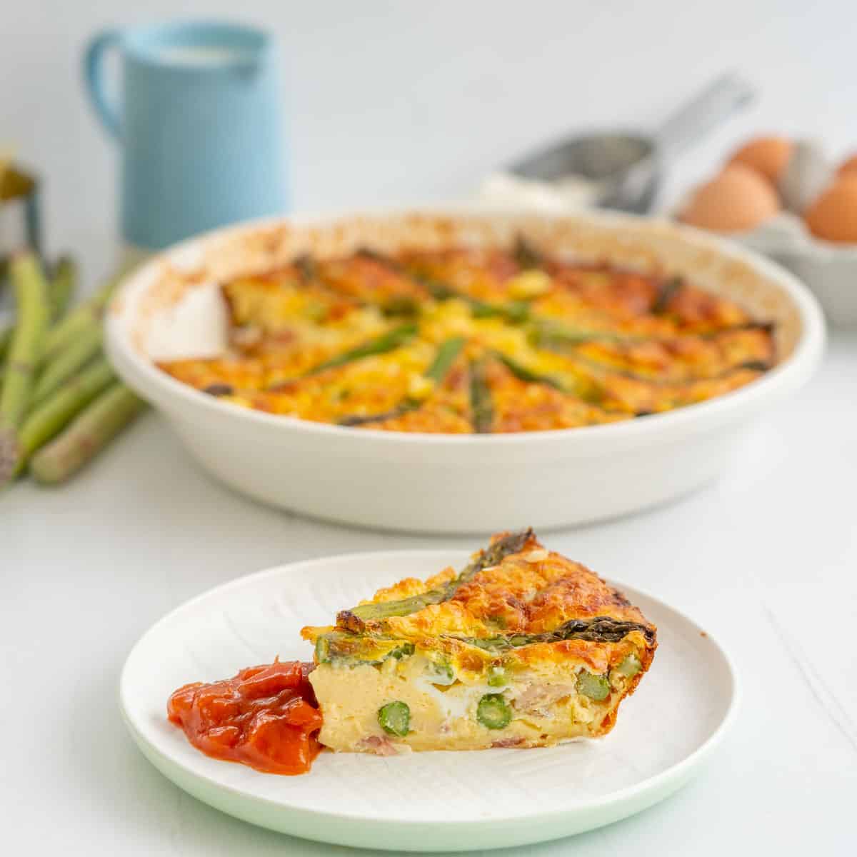 A slice of asparagus quiche  on a white plate garnished with tomato relish, asparagus, a pie dish and eggs in the background. 