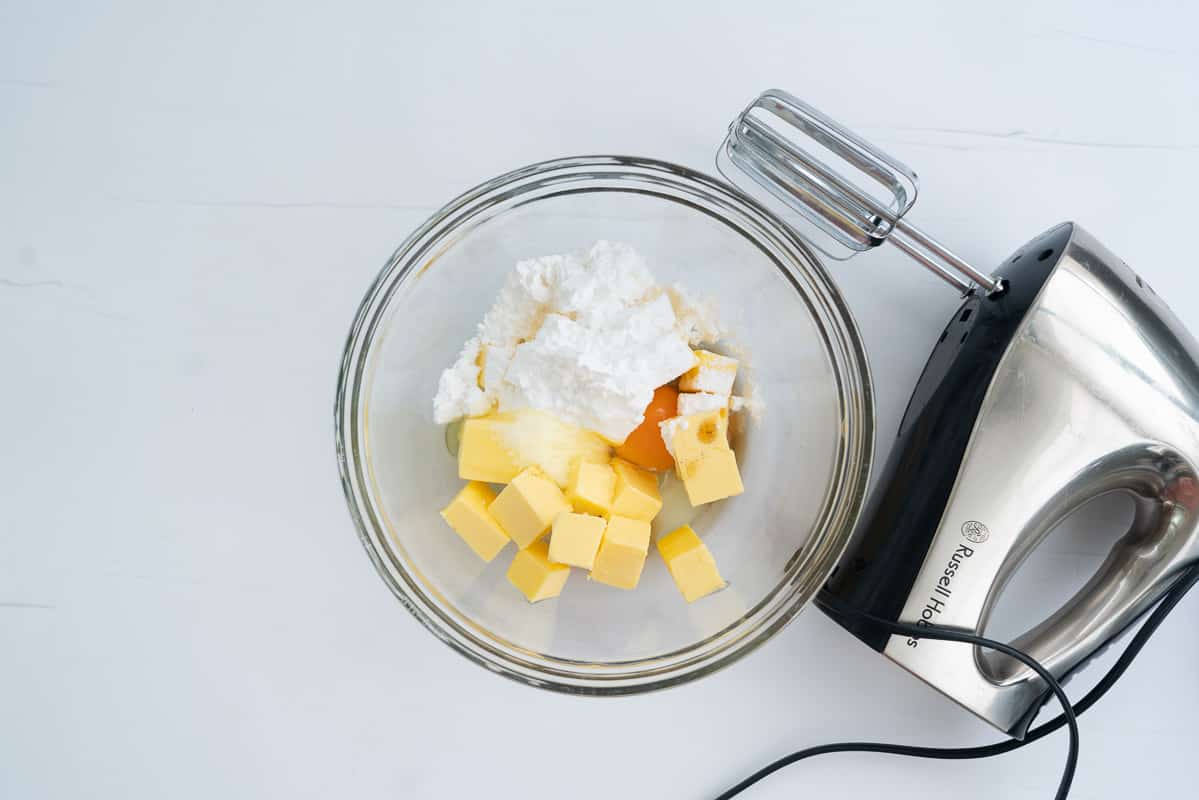 A glass mixing bowl with butter, vanilla essence, eggs and icing sugar in it, next to a hand held electric beater. 