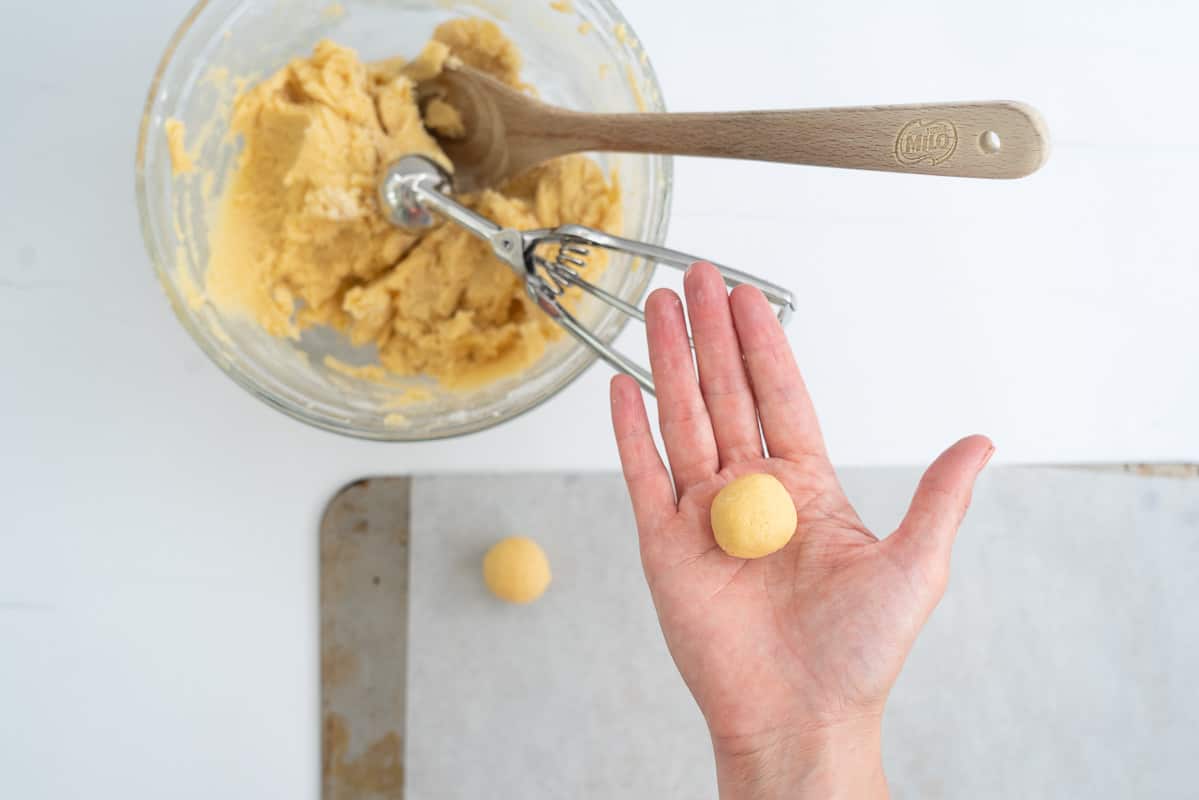 A glass mixing bowl of cookie dough with a cookie scoop, wooden spoon and a hand rolling dough into round balls. 