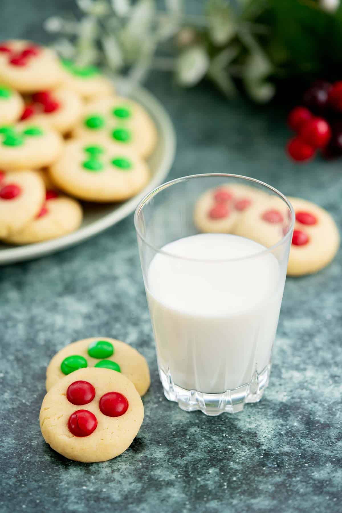 2 cookies decorated with red and green m and ms next to a glass of milk.