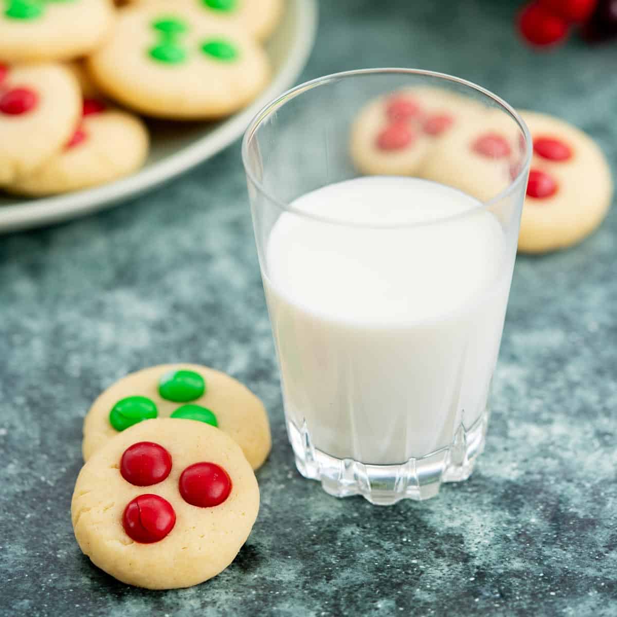 2 cookies decorated with red and green m and ms next to a glass of milk. 