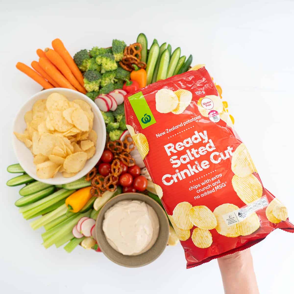A bag of ready salted chips being held above a platter of fresh crudité